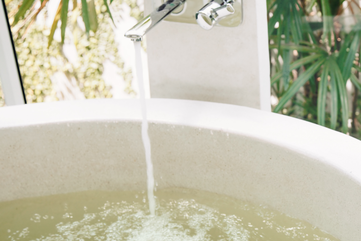 A close-up of water flowing from a chrome faucet into a light beige stone or concrete sink basin, with blurred green foliage visible in the background.